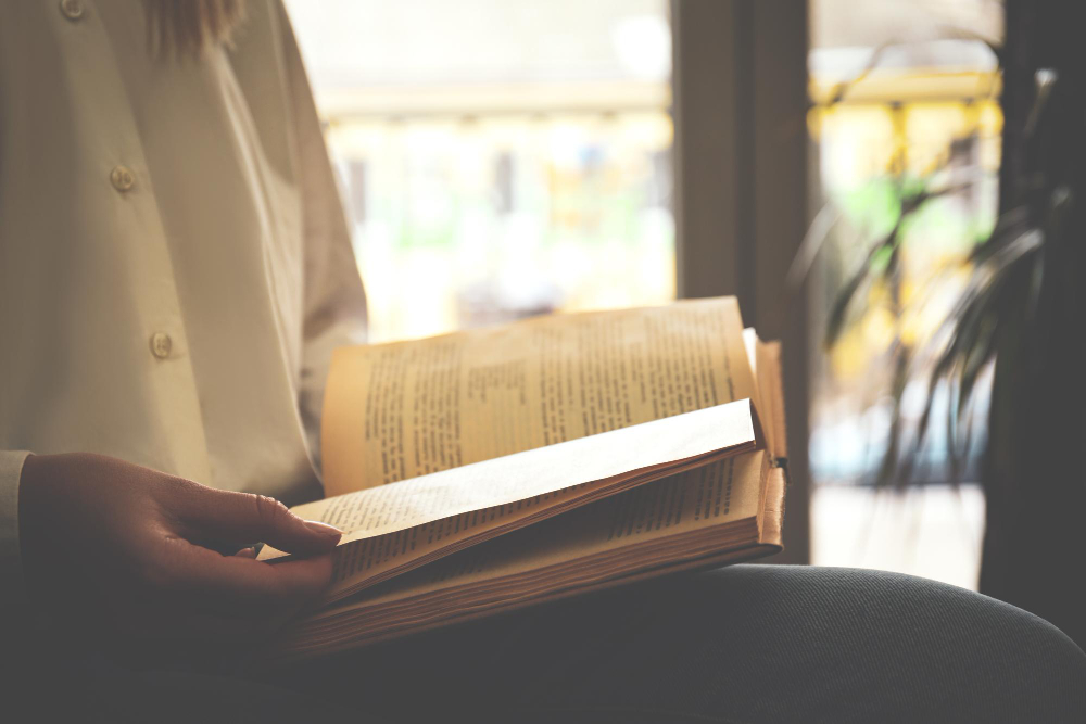 Person in a light shirt reading an open paperback by a sunlit window.
